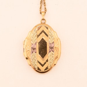May include: An oval gold locket pendant suspended from a matching gold chain. The locket features a central black diamond-shaped inlay, flanked by engraved leaf patterns and small pink floral accents, with chevron-style gold detailing above and below.