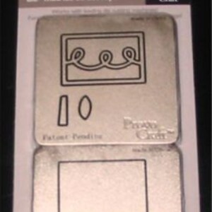 May include: Two metal die cut templates for crafting. The templates are silver and feature a cake design with a wavy top and a cake stand. The text "Provo Craft" is printed on the templates.