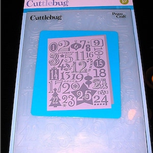 May include: A pink and blue plastic sheet with a gray cutout of numbers and Christmas-themed designs. The sheet is labeled "Cuttlebug" and "Provo Craft".