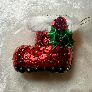 May include: A festive Christmas ornament shaped like a red sequined boot with a white faux fur trim. The boot is adorned with gold accents and a sprig of green holly with red berries. A gold string is attached for hanging.