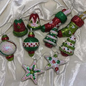 May include: A collection of Christmas ornaments in various shapes, including bells, stockings, Christmas trees, and stars. The ornaments are decorated with red, green, and white sequins and beads, with silver hanging loops.