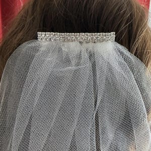 May include: A white tulle veil with a sparkling rhinestone barrette. The barrette is rectangular and features multiple rows of clear rhinestones. The veil is attached to the barrette and drapes down.