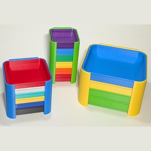 May include: Three sets of square, stackable plastic trays in various colors. The left set has a blue frame with red, white, teal, yellow, blue, and black trays. The center set has a green frame with purple, blue, green, yellow, and red trays. The right set has a yellow frame with blue, yellow, and green trays.