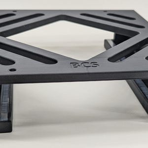 May include: Black plastic 3D printed stand with a square opening and the letters "BCS" printed on the top. The stand has two slots on the sides.