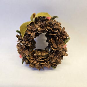 May include: A small, circular wreath composed of brown pinecones. The wreath is adorned with a green ribbon and small pink accents. This decorative wreath is suitable for seasonal home decor.