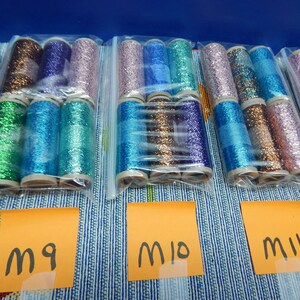 May include: Several spools of colorful glitter thread in clear plastic bags. The thread colors include green, blue, purple, and bronze. Each spool is labeled with a handwritten orange tag. The background is a blue and white striped fabric.