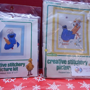 May include: Two Sesame Street creative stitchery picture kits in plastic packaging. One features Cookie Monster with a rolling pin, the other shows Cookie Monster on a seesaw. Finished sizes are 5" x 7" and 9".