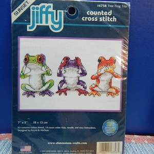 May include: A counted cross stitch kit featuring three colorful tree frogs. The frogs are in shades of green, purple, and orange, each covering their eyes. The kit includes cotton thread, a needle, and instructions. The design measures 7" x 5".