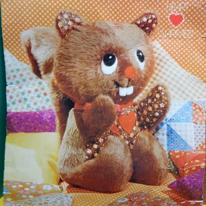 May include: A brown and white stuffed squirrel with a red heart on its chest. The squirrel is sitting on a patchwork quilt. The text "SCAMPER the squirrel HEART WARMER" is printed on the quilt. The text "a springbok stuffed animal kit" is printed on the bottom of the quilt.