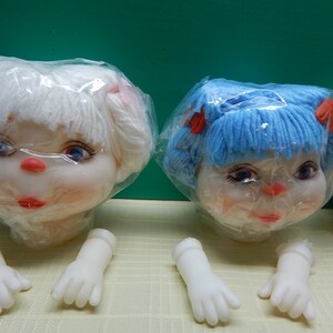 May include: Doll heads with white and blue yarn hair, red bows, and painted faces. The dolls have blue eyes, red lips, and pink cheeks. Detached white plastic doll hands are also visible.