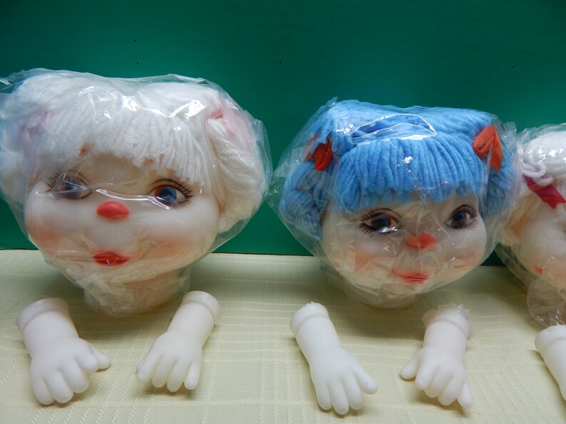 May include: Doll heads with white and blue yarn hair, red bows, and painted faces. The dolls have blue eyes, red lips, and pink cheeks. Detached white plastic doll hands are also visible.