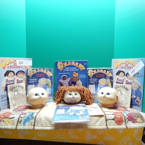 May include: A collection of doll-making supplies, including three doll heads, two books titled "Doll-Baby in Crochet" and "Doll-Baby Easy Clothes to Knit & Crochet", and a book titled "Pre-Sewn Doll Body".