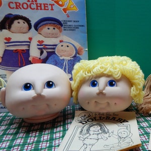 May include: Two doll heads with blue eyes and a vintage crochet pattern book. One doll head is bald, the other has yellow yarn hair. The book cover reads "DOLL-BABY IN CROCHET" with illustrations of crocheted dolls.