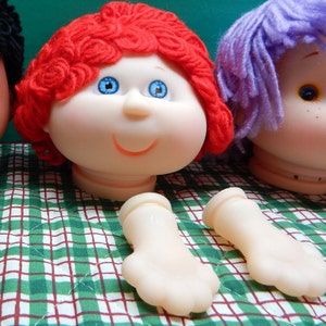 May include: A red yarn doll head with blue eyes and a smile. The doll head is missing its body and has two separate plastic hands.