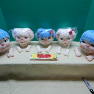 May include: Five vintage doll heads with blue and white yarn hair, each with a smiling face and attached arms. The dolls are arranged on a table, with a "Darice Kids" label visible. The dolls are in plastic wrapping.