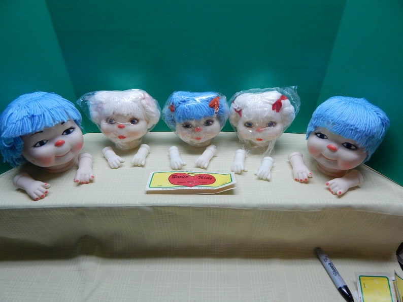 May include: Five vintage doll heads with blue and white yarn hair, each with a smiling face and attached arms. The dolls are arranged on a table, with a "Darice Kids" label visible. The dolls are in plastic wrapping.