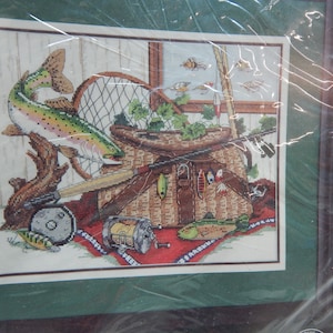 May include: Cross-stitch artwork depicting a fishing scene. A rainbow trout leaps above a woven basket filled with fishing gear, including lures and a fishing rod. The background shows a window with insects and a wooden wall.