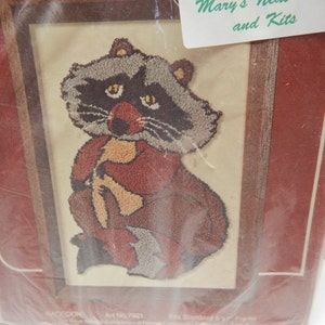 May include: A framed raccoon needlepoint kit. The raccoon is depicted in shades of brown, gray, and red, holding a yellow object. The frame is a dark wood color. The kit is in a plastic sleeve, with the text "Mary's Needle and Kits" visible.