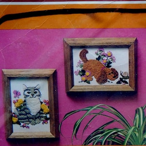 May include: Two framed embroidery pieces, one with a cat and the other with a dog, both surrounded by flowers. The frames are wooden with a light brown finish. The background is a bright pink.