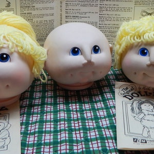 May include: Three doll heads with blonde yarn hair and blue eyes. One head is bald. The doll heads are on a green and white checkered surface. Two copies of a vintage "The Original DOLL-BABY" pattern are in the foreground.