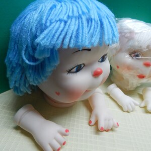 May include: A doll head with blue yarn hair, rosy cheeks, and a red nose. The doll has blue eyes and a small red heart on its cheek. Detached hands and feet with painted red fingernails are also visible.