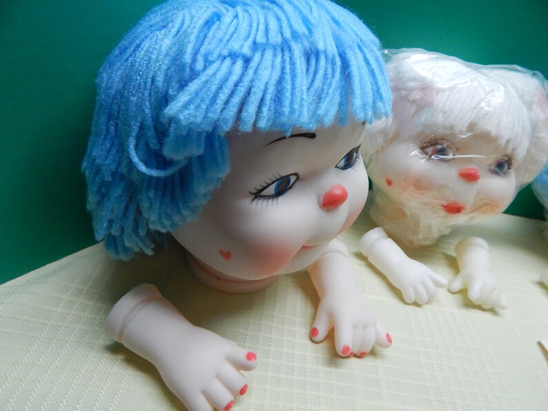 May include: A doll head with blue yarn hair, rosy cheeks, and a red nose. The doll has blue eyes and a small red heart on its cheek. Detached hands and feet with painted red fingernails are also visible.