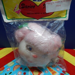 May include: A doll head in a clear plastic bag. The doll has a pale complexion, blue eyes, and pink yarn hair. The packaging includes the text "Darice DOLLS WITH A HEART" and "Mary's Neat Kits and More".
