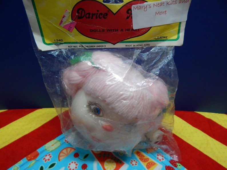 May include: A doll head in a clear plastic bag. The doll has a pale complexion, blue eyes, and pink yarn hair. The packaging includes the text "Darice DOLLS WITH A HEART" and "Mary's Neat Kits and More".
