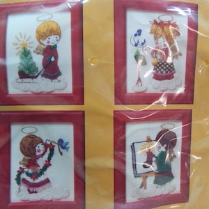 May include: Four framed embroidered pictures of angels. Each angel has a halo and wings, and is depicted in different scenes, such as with a Christmas tree or painting. The frames are red, and the background is white.