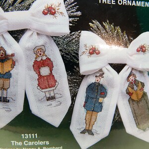 May include: White fabric Christmas tree ornaments with cross-stitched carolers. Each ornament has a bow at the top with embroidered red berries. The carolers are dressed in winter clothing. The text "The Carolers" is visible.