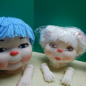 May include: Doll heads with blue and white yarn hair, red noses, and painted facial features. The dolls have detached arms and hands. The background is a solid green color.