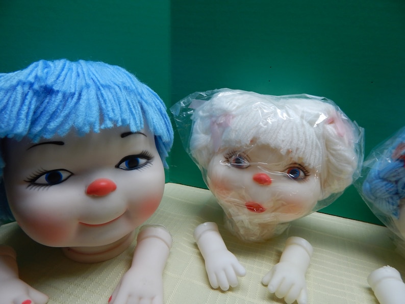 May include: Doll heads with blue and white yarn hair, red noses, and painted facial features. The dolls have detached arms and hands. The background is a solid green color.