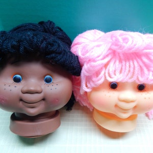 May include: Two doll heads with yarn hair. The left doll has dark skin, black yarn hair, blue eyes, and freckles. The right doll has light skin, pink yarn hair, blue eyes, and freckles. Both dolls have a smiling expression and rosy cheeks.