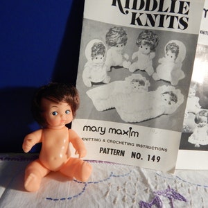 May include: A plastic doll with brown hair and blue eyes, sitting on a white surface. The doll is wearing no clothes. A black and white knitting pattern booklet is in the background. The booklet is titled "Riddle Knits" and "Mary Maxim Knitting & Crocheting Instructions, Pattern No. 149".
