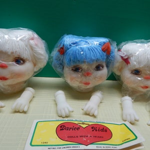May include: Three doll heads with yarn hair in white and blue, each in a plastic bag. The dolls have painted faces with blue eyes and red lips. Detached white plastic hands are below. A Darice Kids label is visible.