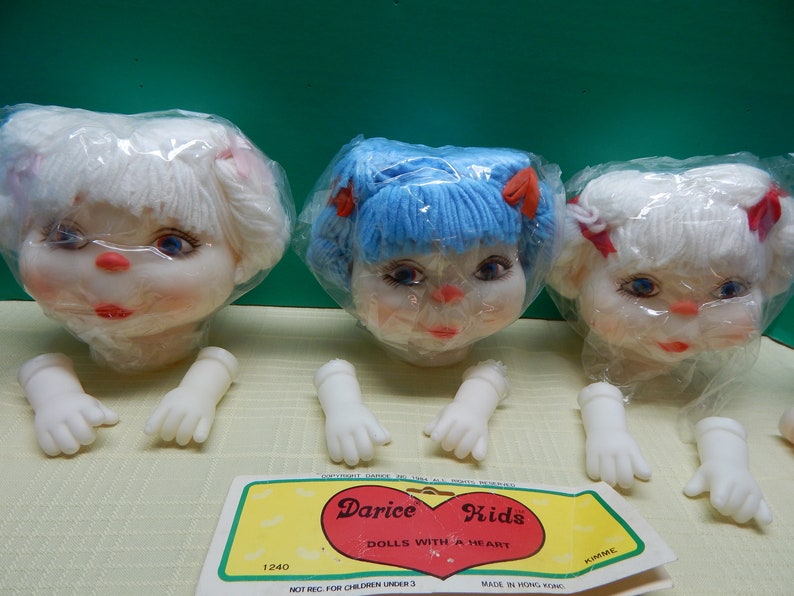 May include: Three doll heads with yarn hair in white and blue, each in a plastic bag. The dolls have painted faces with blue eyes and red lips. Detached white plastic hands are below. A Darice Kids label is visible.