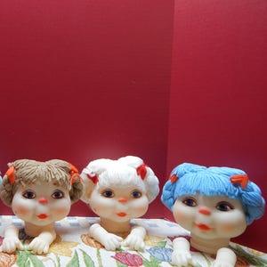 May include: Three doll heads with different colored yarn hair, each with a red bow. The dolls have painted faces with blue eyes and red lips. They are resting on a floral patterned fabric.