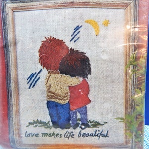 May include: Embroidered artwork in a wooden frame depicts two figures embracing, with text reading "love makes life beautiful." The figures have red and brown hair, wearing blue jeans and a red coat. A yellow crescent moon and star are in the sky.