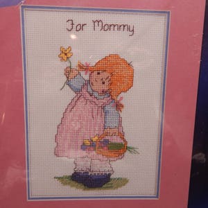 May include: Cross-stitch embroidery of a girl with red hair holding a basket of flowers. The text "For Mommy" is above the girl.