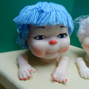 May include: A doll head with blue yarn hair, rosy cheeks, and a red nose. The doll has blue eyes and is smiling. Detached arms and hands with red painted fingernails are also visible. The doll is on a yellow surface.