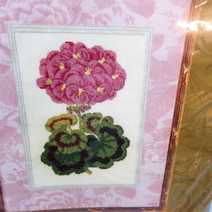 May include: Cross-stitch embroidery of a pink geranium flower with green leaves, framed in a wooden frame. The background is a light pink floral pattern. The embroidery is detailed and colorful, showcasing the flower's petals and leaves.