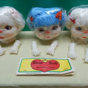 May include: Three doll heads with white, blue, and white yarn hair, each in a plastic bag. White plastic doll hands are arranged below. A yellow and green label reads "Darice Kids DOLLS WITH A HEART".
