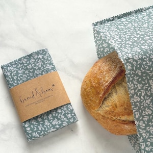 May include: A loaf of bread partially wrapped in a teal floral fabric bread bag. Beside it is a matching folded bread wrap with a brown paper label that reads "bread & bees". The bag and wrap are made of fabric with a white floral pattern.