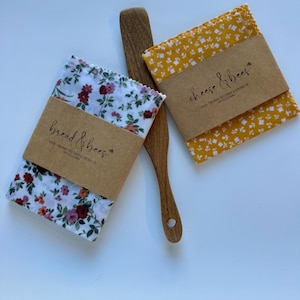 May include: Two fabric food wraps with floral patterns and brown paper labels that read "bread & bees" and "cheese & bees". A wooden spatula is placed between the wraps. The wraps are white with red and purple flowers and yellow with white flowers.