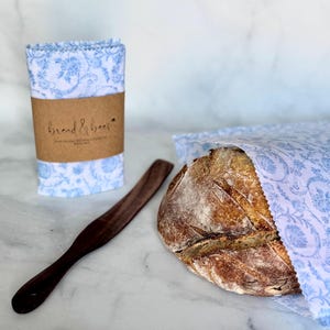 May include: A loaf of bread wrapped in a blue and white floral patterned cloth, next to a stack of matching cloths and a wooden bread knife. The stack of cloths is wrapped with a brown paper band that reads "bread & bees".