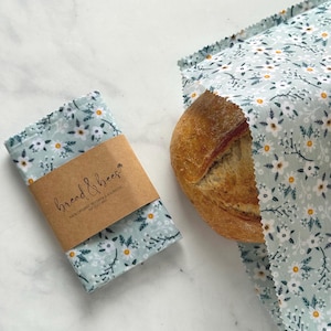 May include: A loaf of bread partially wrapped in a light blue floral fabric bread bag. A matching folded bread wrap with a brown paper label that reads "bread & bees" is next to the bread. The fabric has a white daisy pattern.