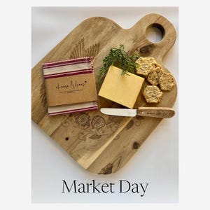 May include: A wooden cheese board with a handle, featuring a block of cheddar cheese, crackers, a cheese knife, and a small package wrapped in brown paper and red striped fabric. The text "cheese & bees" is visible on the package, and the text "Market Day" is at the bottom.