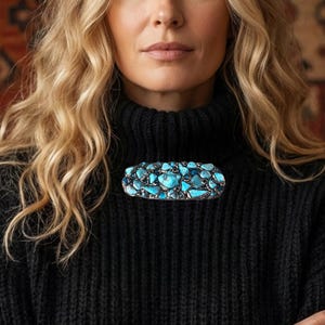May include: A striking turquoise and silver brooch, oval in shape and studded with turquoise stones, is the central feature. The person is wearing a black turtleneck jumper, complemented by silver rings and bracelets.