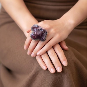 May include: A large statement ring featuring a raw, deep purple amethyst crystal cluster set in a textured silver-toned metal band. The stone has an irregular, jagged surface with natural crystalline facets and dark violet hues throughout the piece.