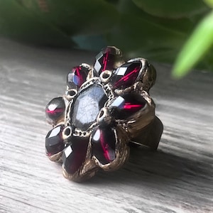 May include: A vintage-style ring with a floral design. The ring features a central dark stone surrounded by faceted, deep red gemstones. The setting is a gold-toned metal, and the ring is displayed on a wooden surface.